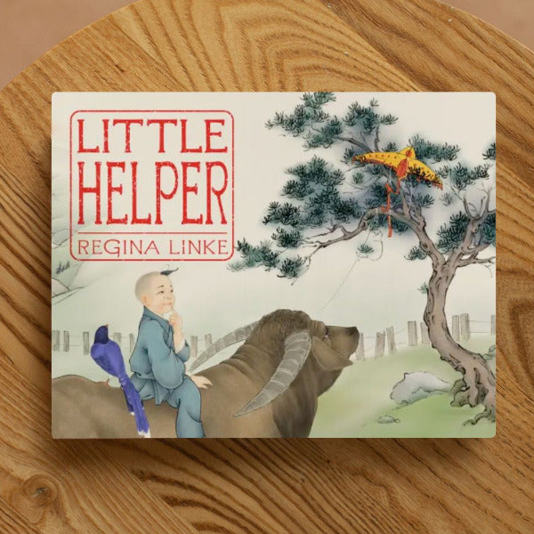 Book titled Little Helper by Regina Linke on a wood table shows a Chinese painting of a boy and a Taiwanese magpie perched on the back of an ox, looking at a kite stuck in a tree in the background.