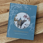 The Oxherd Boy book in English with a blue cover and lighter blue bamboo frame an inset circle displaying a Chinese art painting of a young boy in blue riding a brown ox with a white rabbit behind him. The background shows a peaceful countryside and farmhouse with an orange roof, flanked by two trees, one with red blossoms, the other with yellow leaves